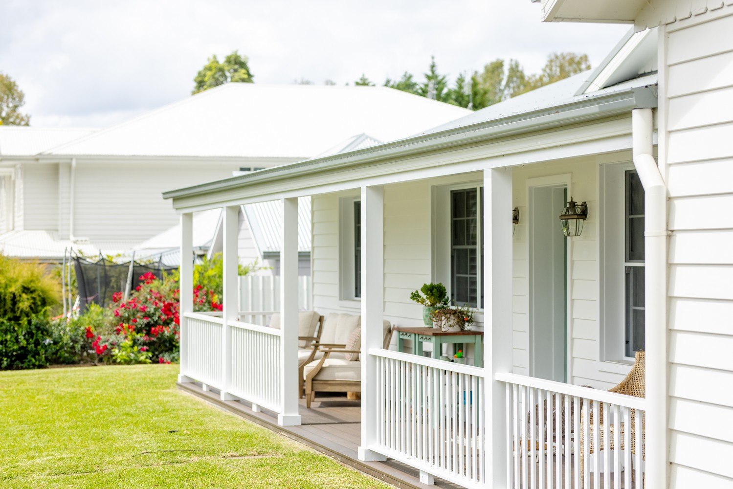 A charming white house with a veranda, featuring comfortable seating and greenery, surrounded by vibrant flowers in the garden.