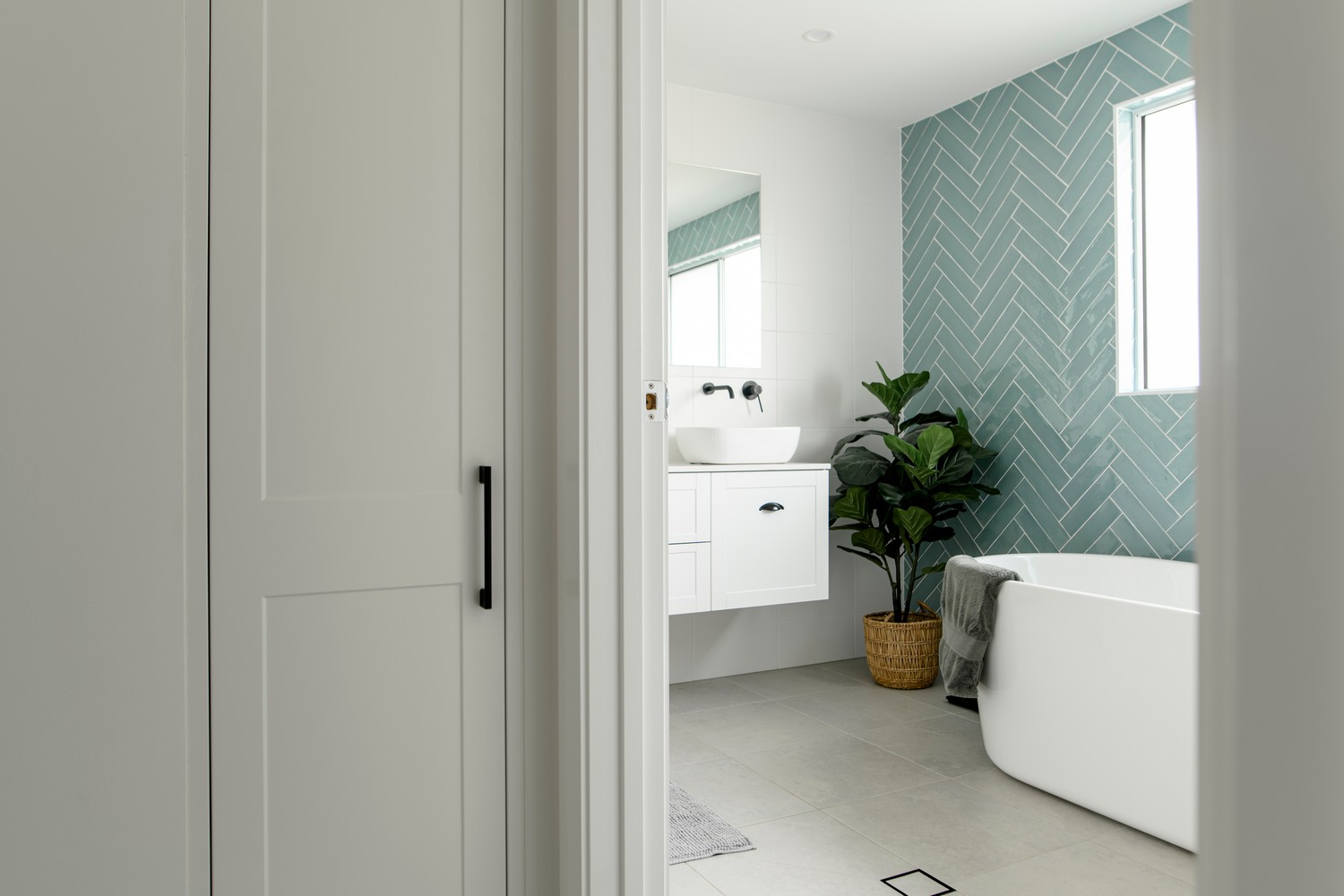 Modern bathroom featuring a white tub, herringbone-patterned teal wall tiles, a sleek vanity, and a potted plant. Bright and inviting space.