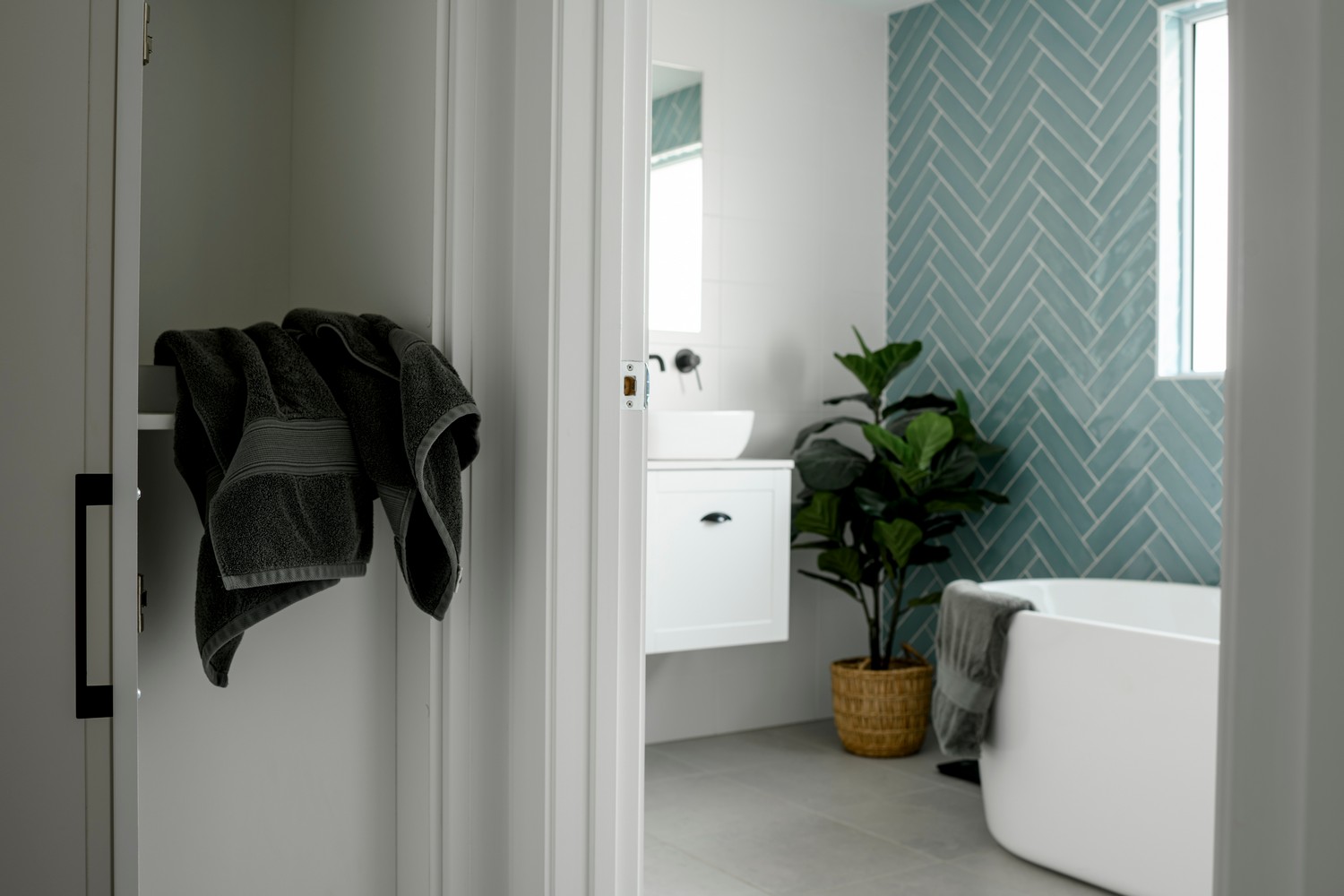 A modern bathroom featuring a white bathtub, green herringbone tiles, a potted plant, and grey towels hanging on the wall.