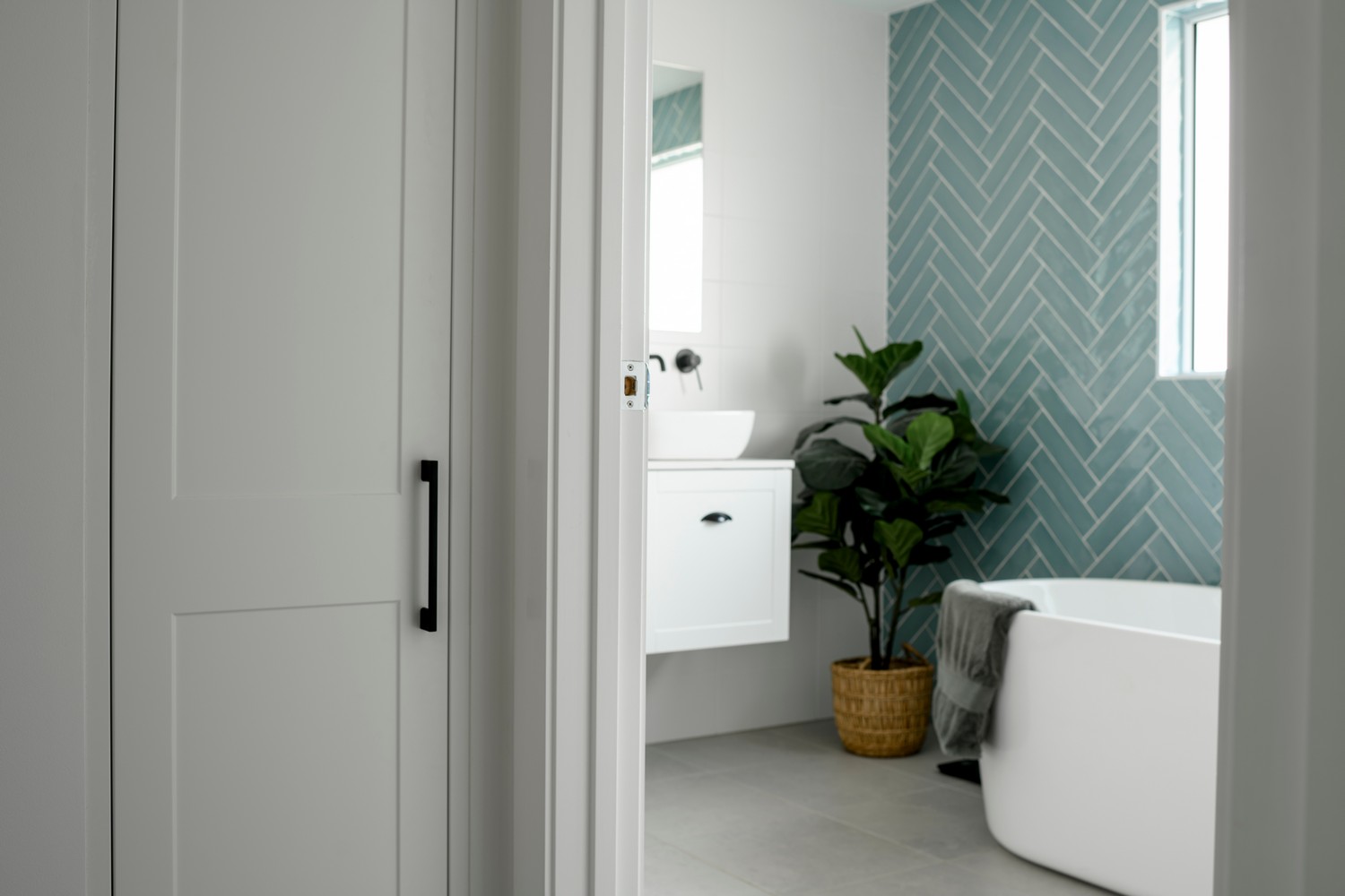 A modern bathroom with a white bath, teal herringbone tile wall, potted plant, and sleek cabinetry, viewed from a doorway.