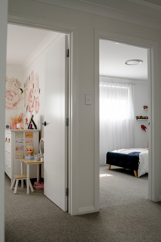 Brightly lit hallway leading to a softly decorated bedroom. Floral wallpaper, a white dresser, and a bed with a dark blue cover create a cosy atmosphere.