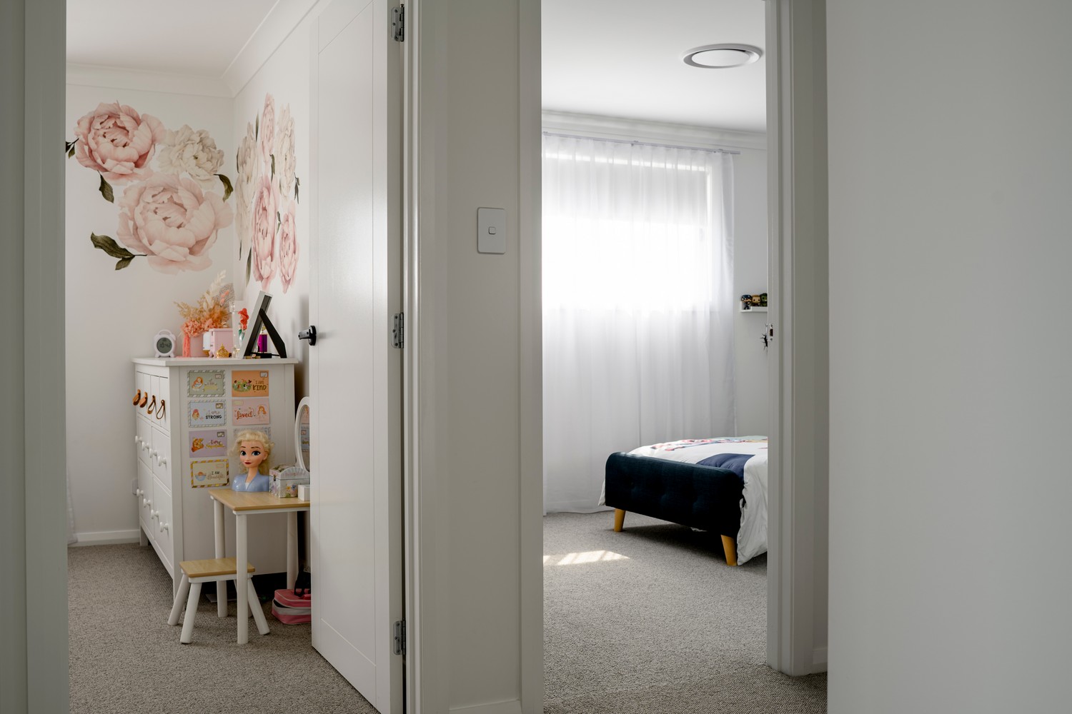 Bright and inviting room featuring a floral wall mural, light-coloured walls, beige carpet, and a view into a neatly arranged bedroom.
