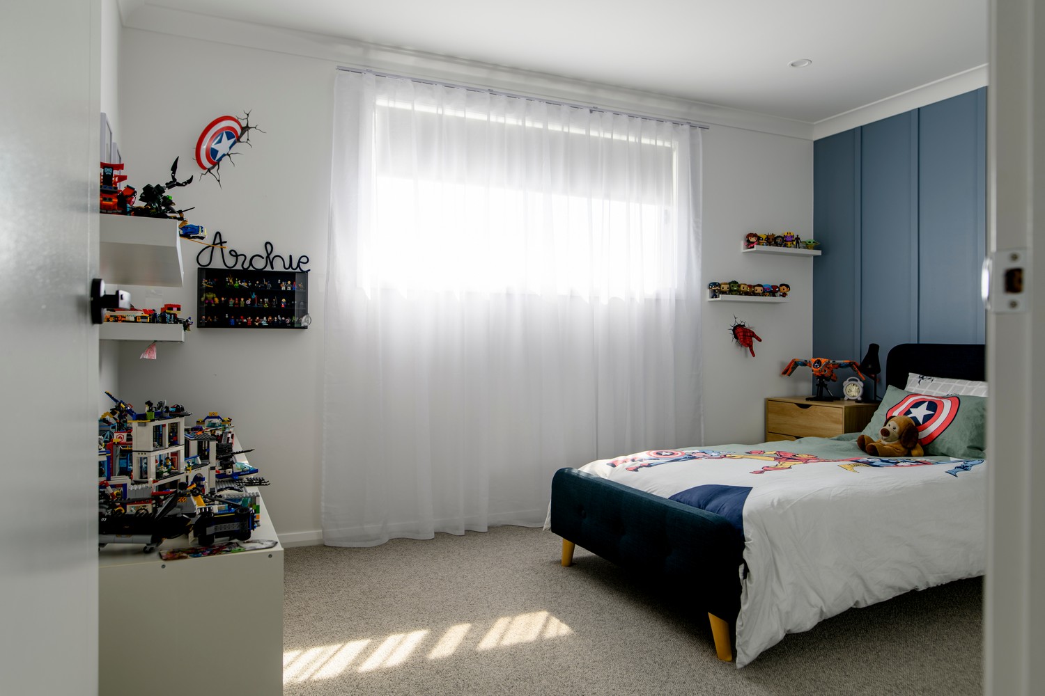 Bright children's bedroom featuring a blue accent wall, a bed with superhero bedding, and shelves displaying toy figurines.