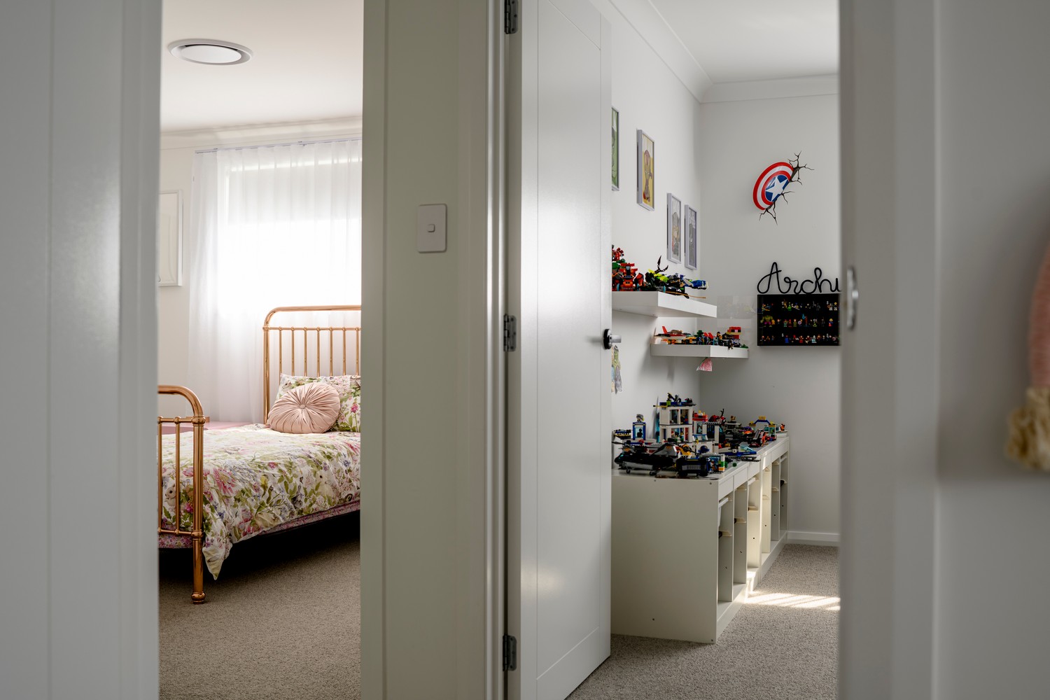 Brightly lit children's bedroom featuring a pink metal bed with floral bedding and a toy display unit filled with colourful toys.