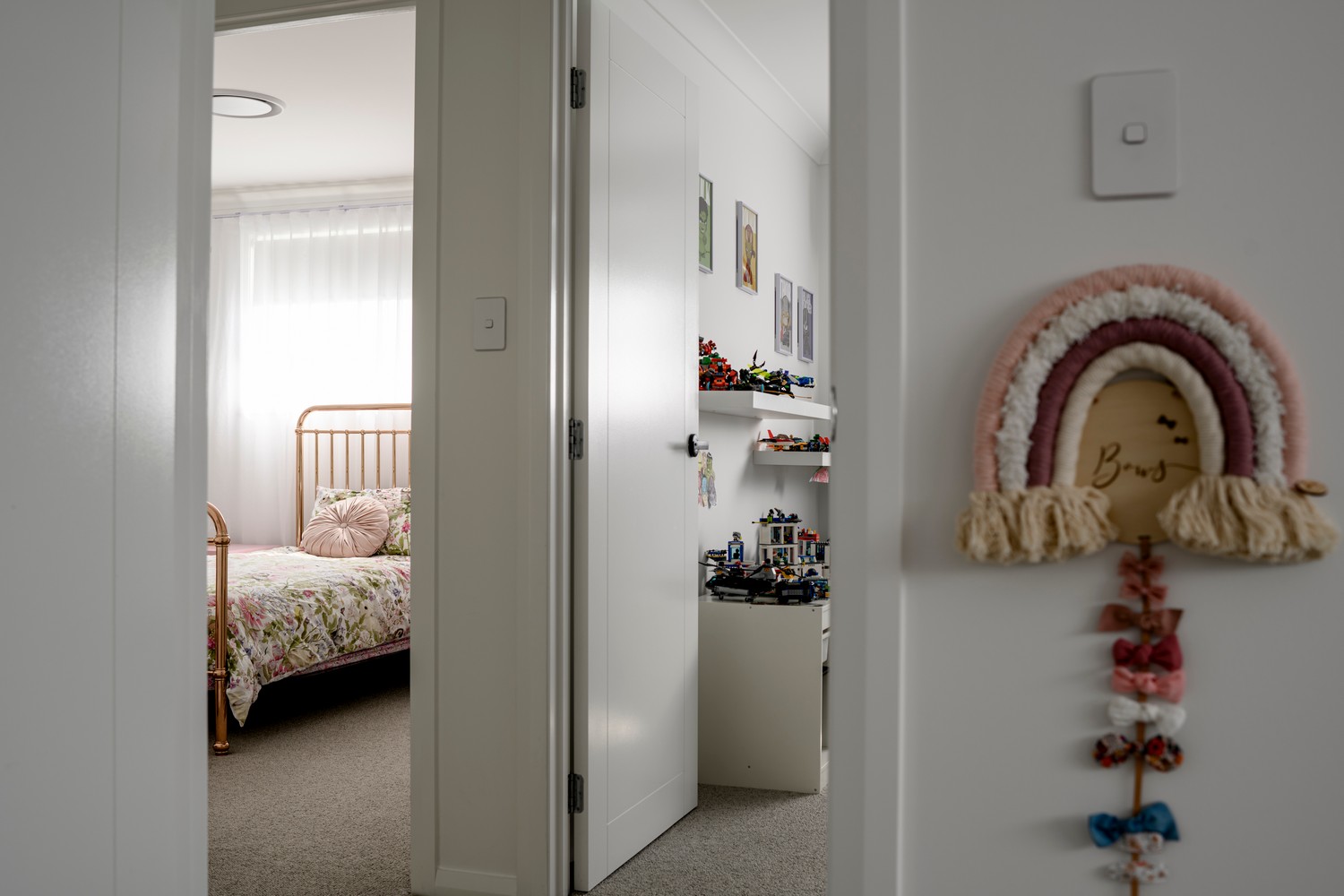 A softly lit hallway leads to a decorated child's bedroom with floral bedding, toys on shelves, and a rainbow wall hanging nearby.