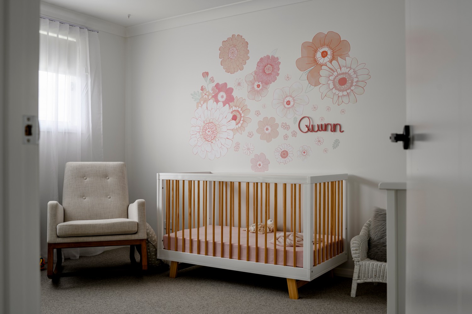 A softly lit nursery featuring a white crib with a peach-coloured interior, a cream armchair, and floral wall decals in pink hues.