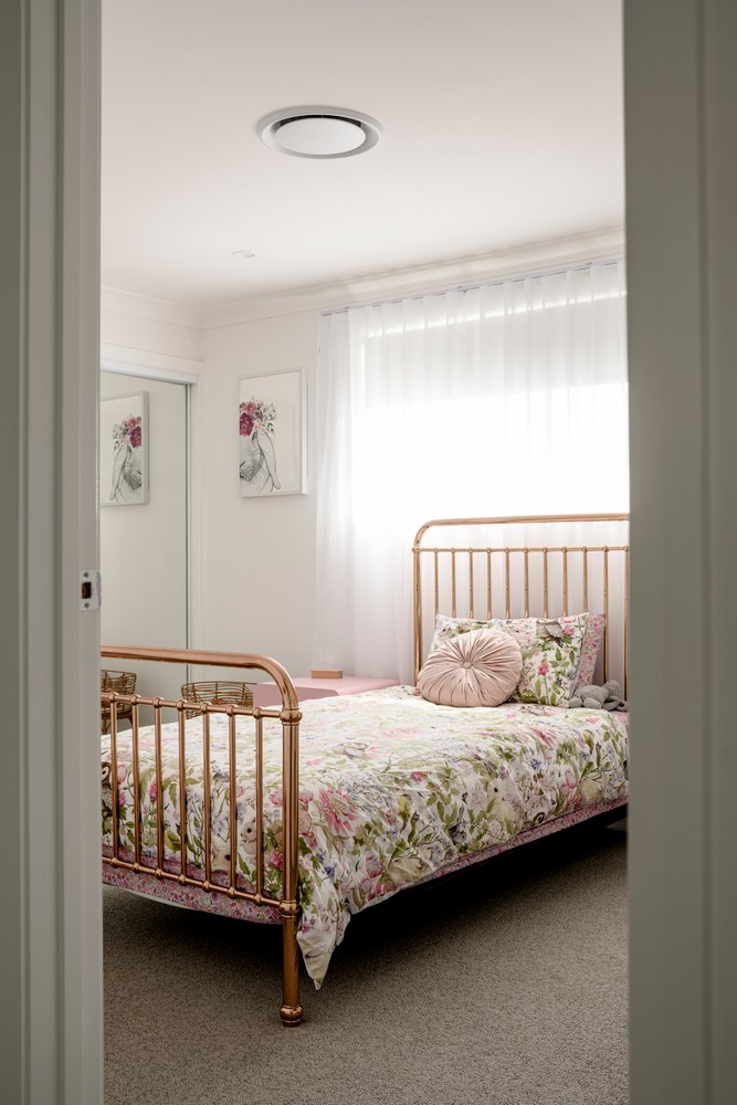 A softly lit bedroom featuring a rose gold bed with a floral quilt, plush cushions, and light curtains, creating a serene atmosphere.