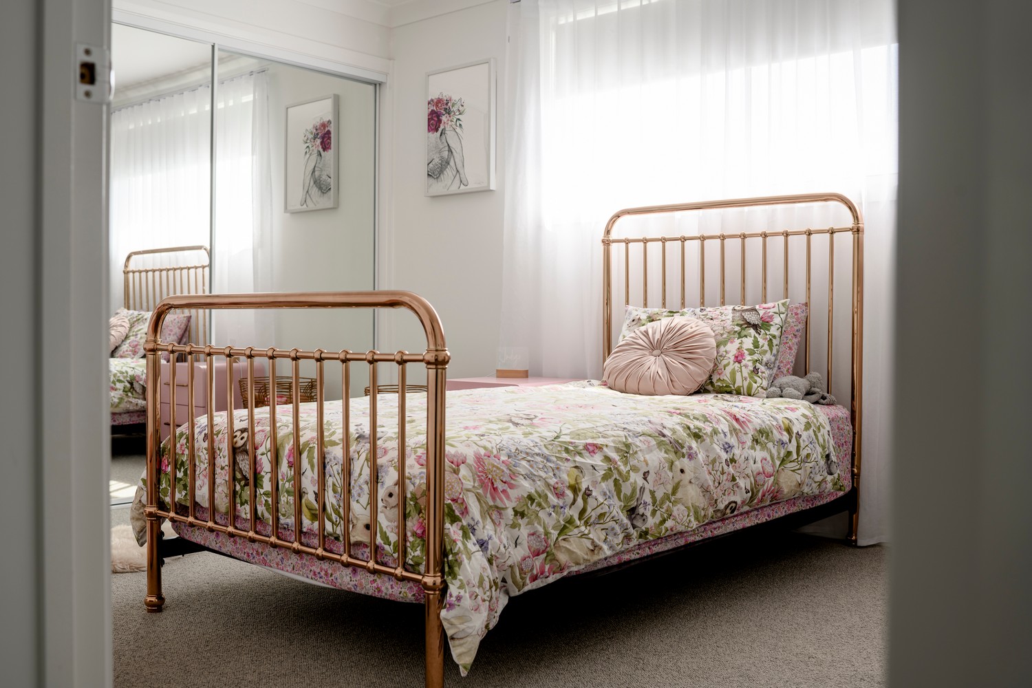 A beautifully styled bedroom featuring a rose gold bed frame, floral bedding, and large mirrors reflecting soft, natural light.