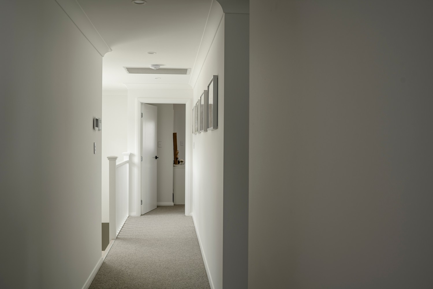 Bright, minimalist hallway with light walls, a beige carpet, and framed pictures, leading to a doorway at the far end.