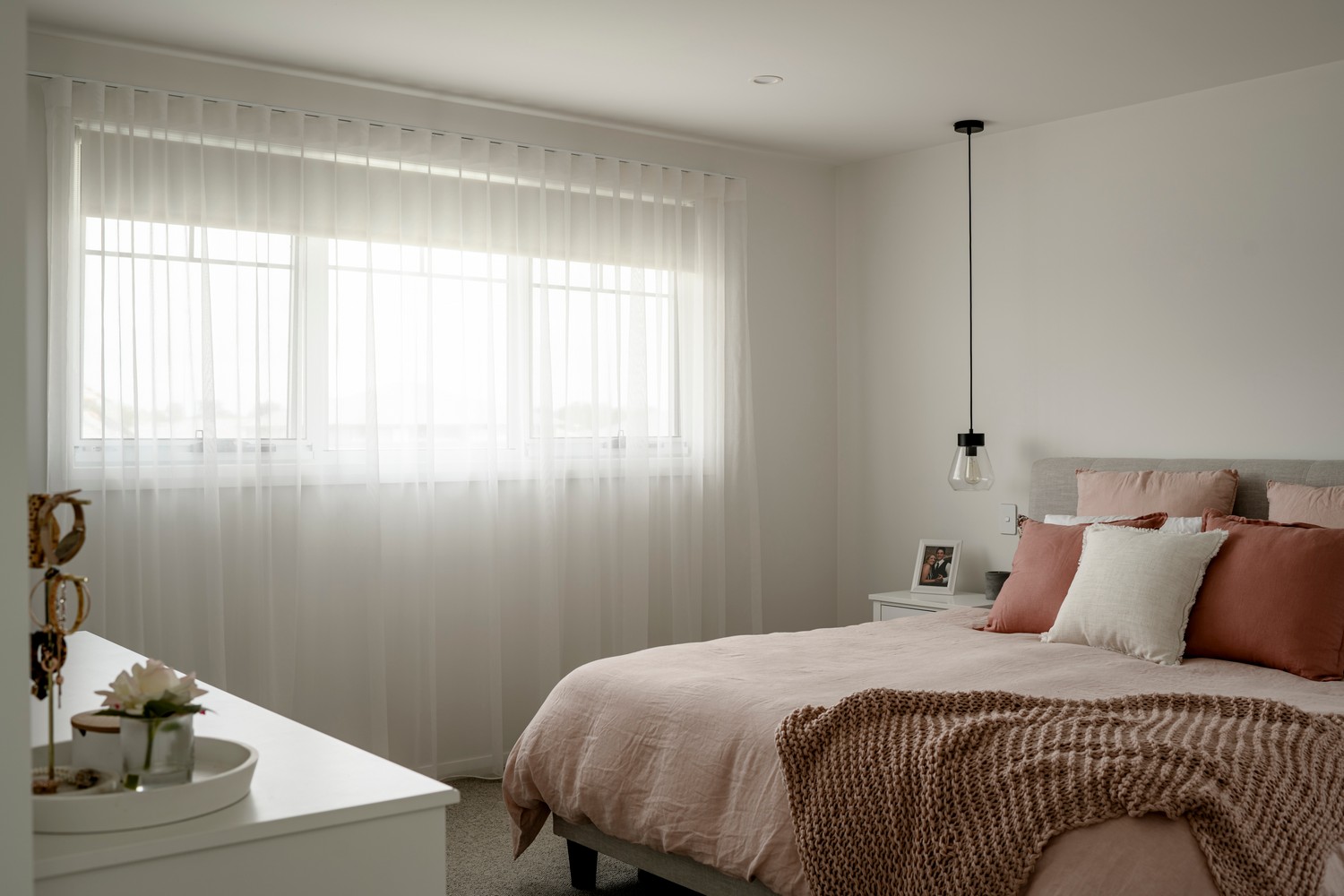 A serene bedroom featuring a soft pink bedspread, textured cushions, and sheer white curtains, creating a warm and inviting atmosphere.