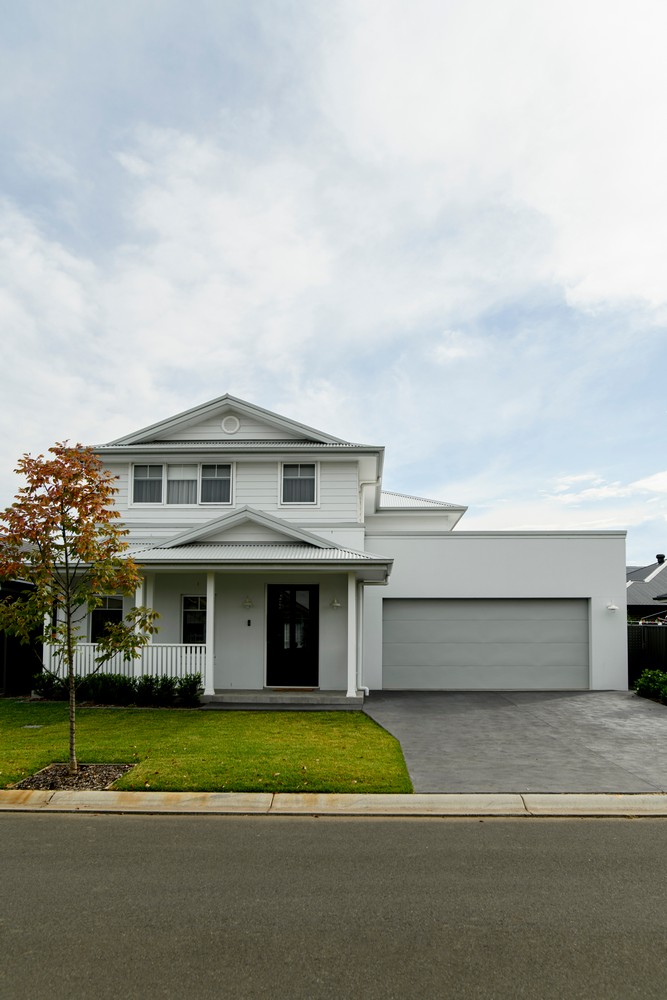 A modern two-storey house with a white facade, black door, and garage, surrounded by green grass and trees under a cloudy sky.