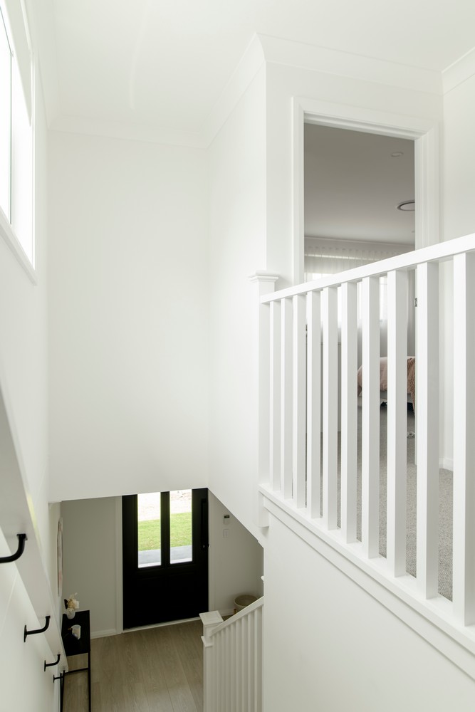 Bright, minimalist staircase leading to an upper floor, with white walls, wooden flooring, and a dark front door visible at the bottom.