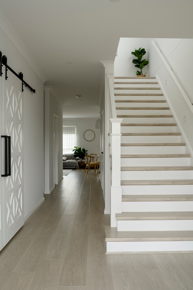 Bright hallway with a wooden staircase, light walls, a sliding barn door, and a potted plant, leading to a living area with natural light.