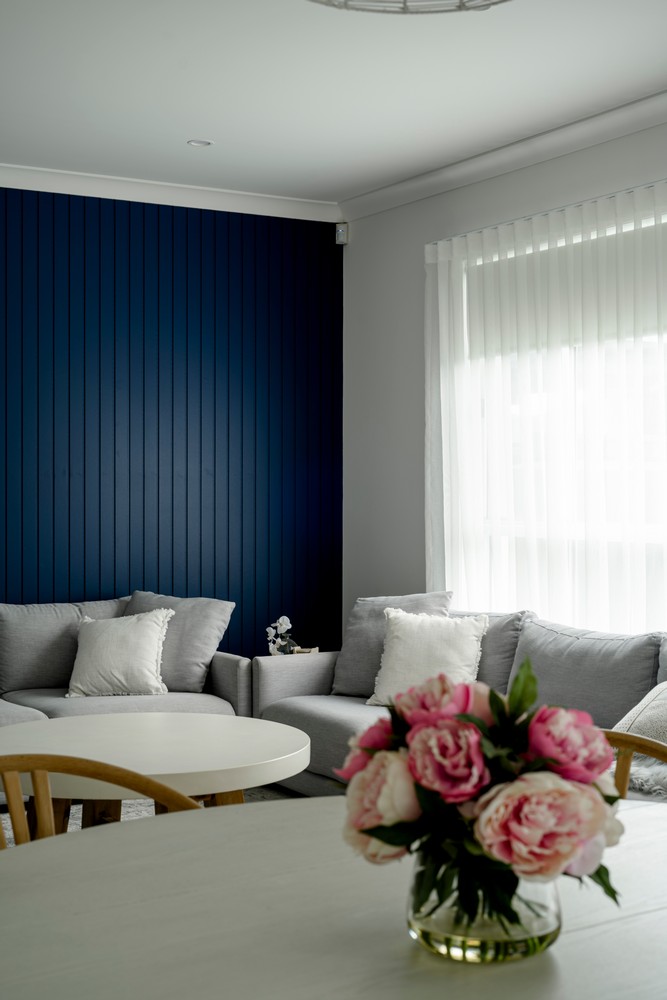 Bright living space featuring a navy blue accent wall, grey sofas with cushions, a light coffee table, and a vase of pink peonies.