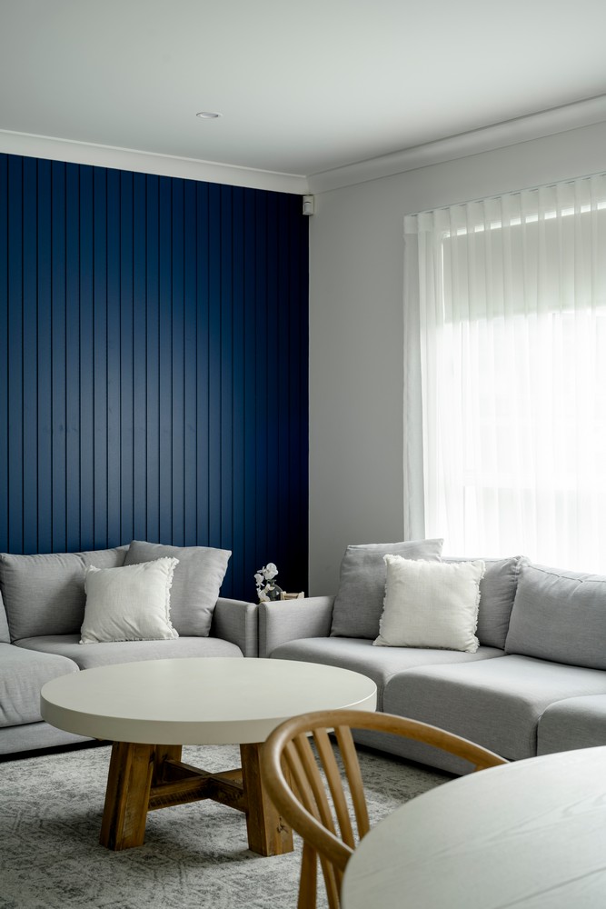 A modern living room with a navy blue feature wall, light grey sofas, a round white table, and sheer curtains letting in natural light.