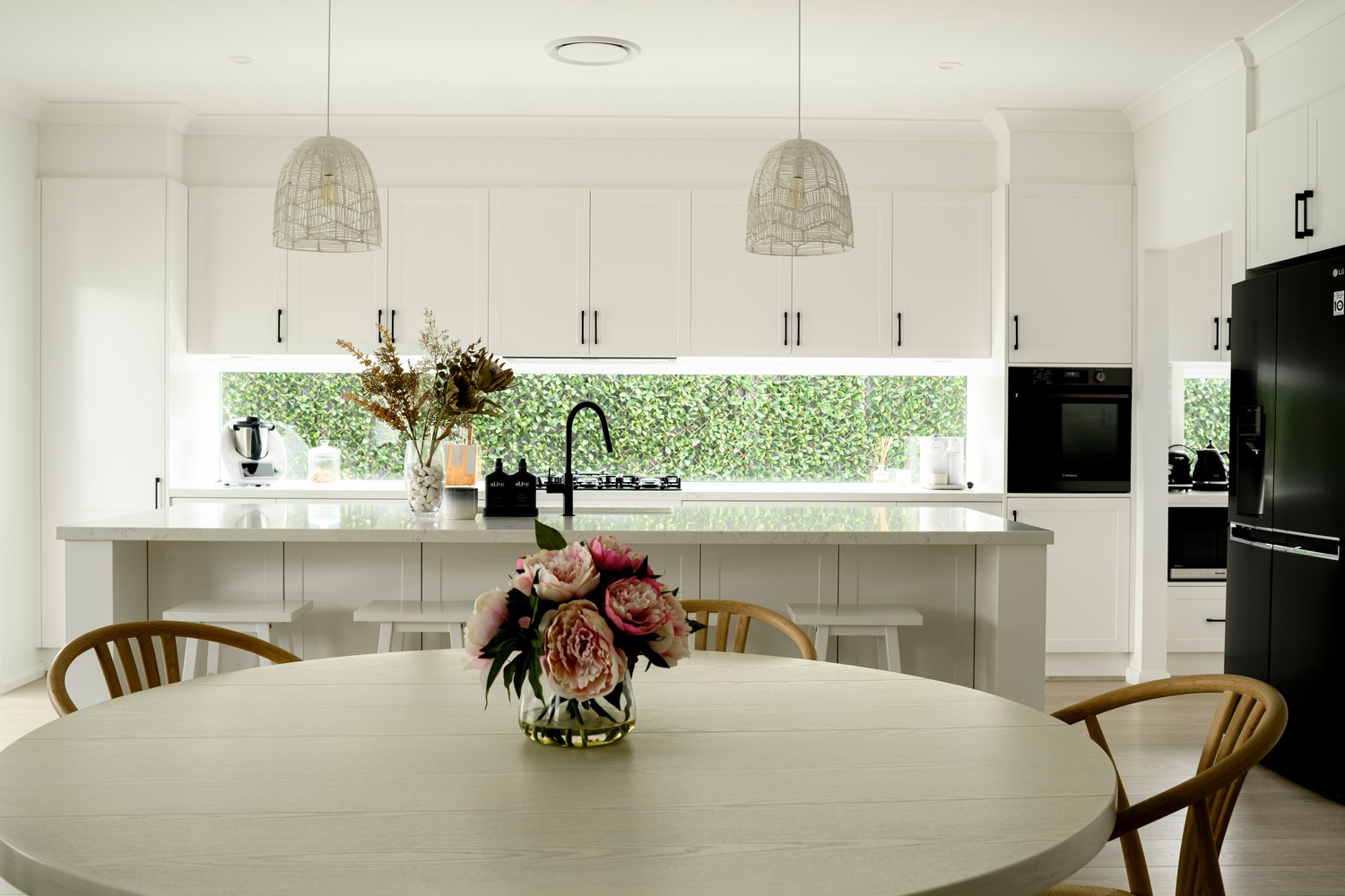 Modern kitchen featuring white cabinetry, a central island, and green foliage in the background. A vase of pink flowers adorns a round table.