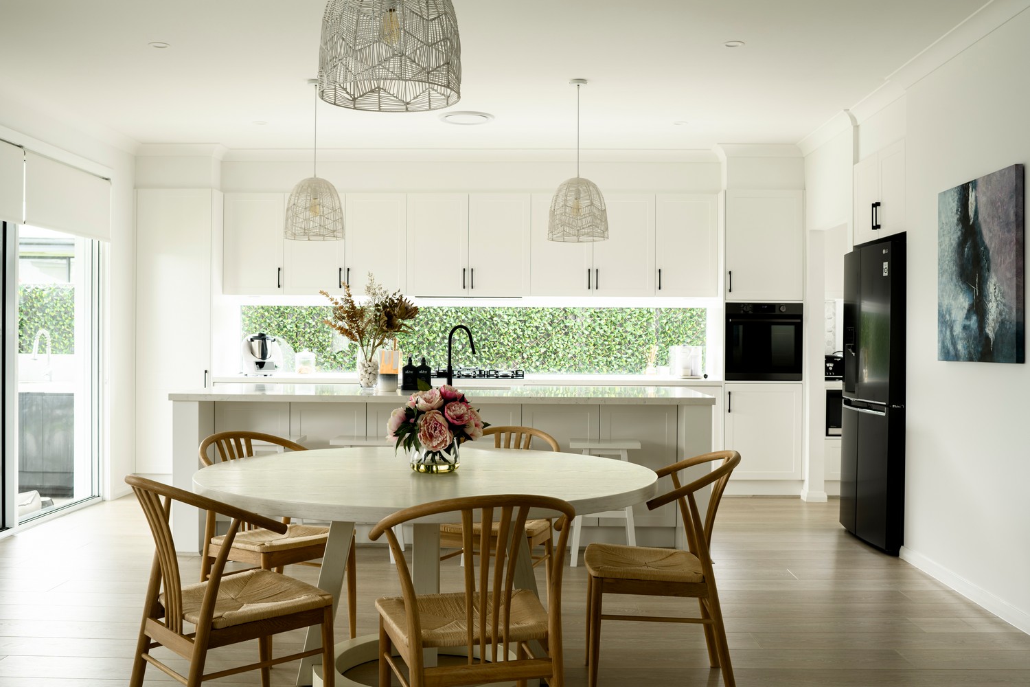Bright, modern kitchen with white cabinetry, a central circular table, wooden chairs, and greenery visible through large windows.