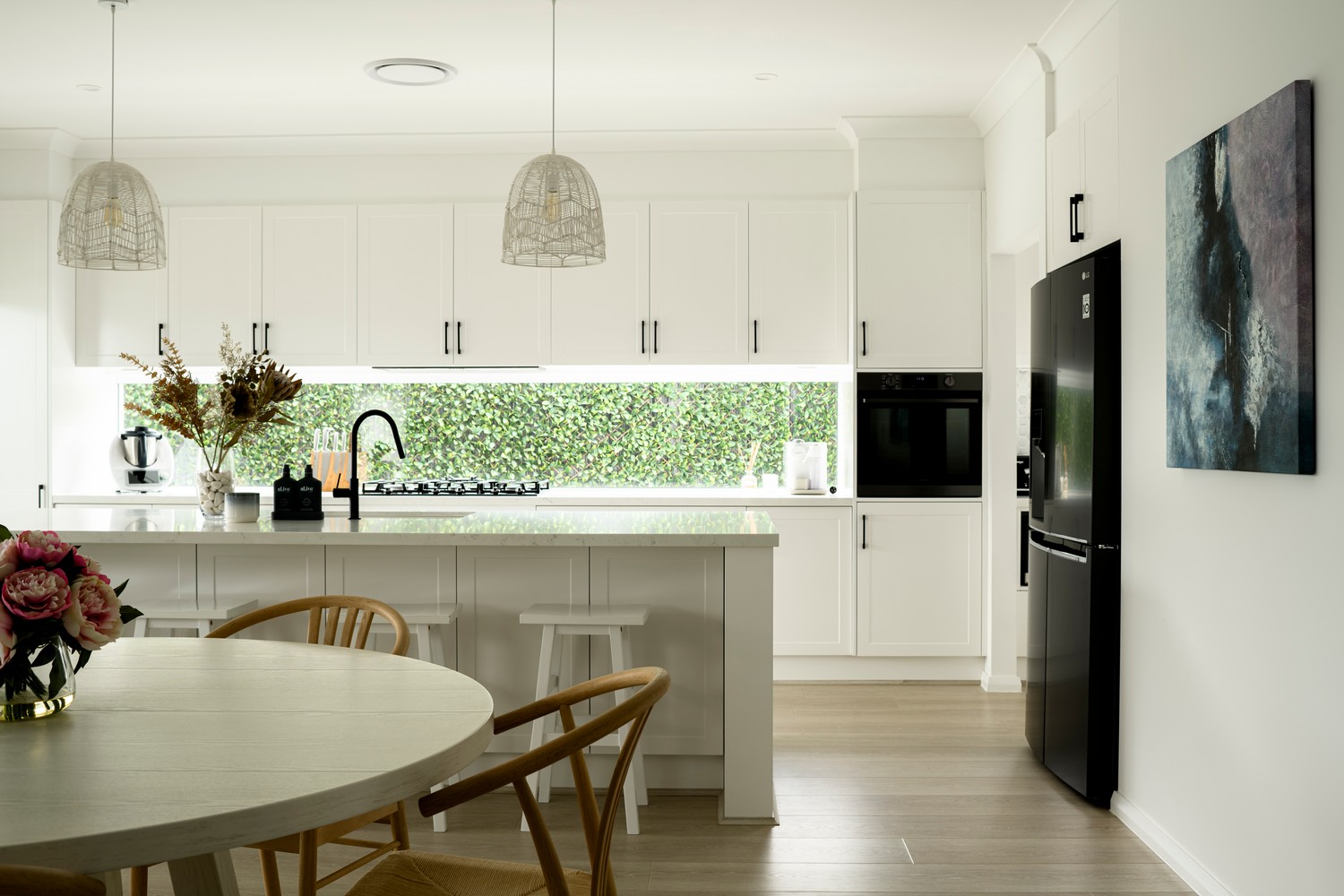 Bright and modern kitchen featuring white cabinetry, a large island, black appliances, and greenery visible through a window, complemented by subtle decor.