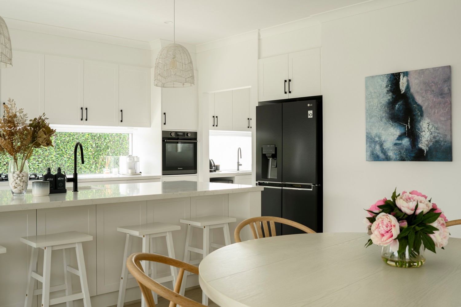 Modern kitchen featuring white cabinetry, black appliances, a round dining table with a floral arrangement, and stylish pendant lighting.