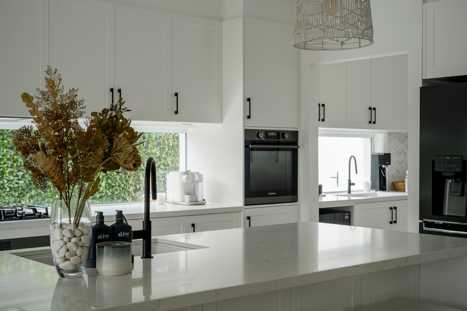 Bright modern kitchen featuring white cabinetry, black appliances, a large island with a marble countertop, and a vase of dried flowers.