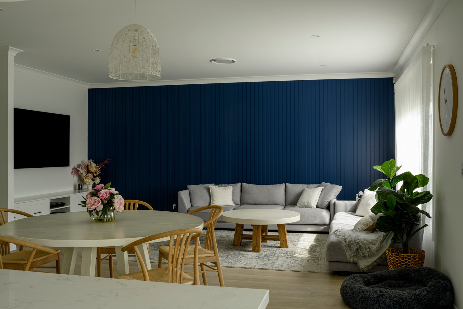 Bright living space featuring a navy blue accent wall, grey sofa, round white coffee table, and a dining area with wooden chairs.