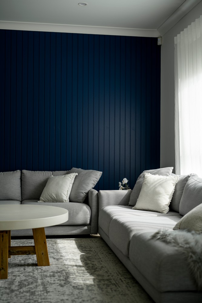 A modern living room features a grey sectional sofa with cushions, a round white coffee table, and a navy blue panelled wall.
