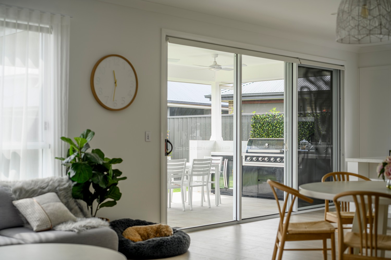 Bright, modern interior featuring a sliding glass door leading to an outdoor area with a barbecue, dining table, and greenery, complemented by a clock and indoor plants.