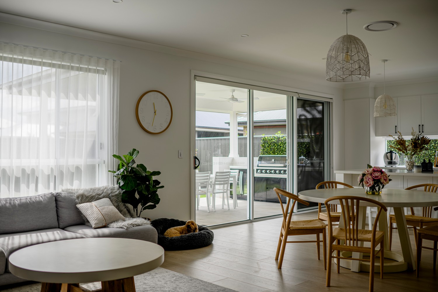 Bright, modern interior with a grey sofa, round coffee table, and wooden dining set. Natural light fills the space through large glass doors.