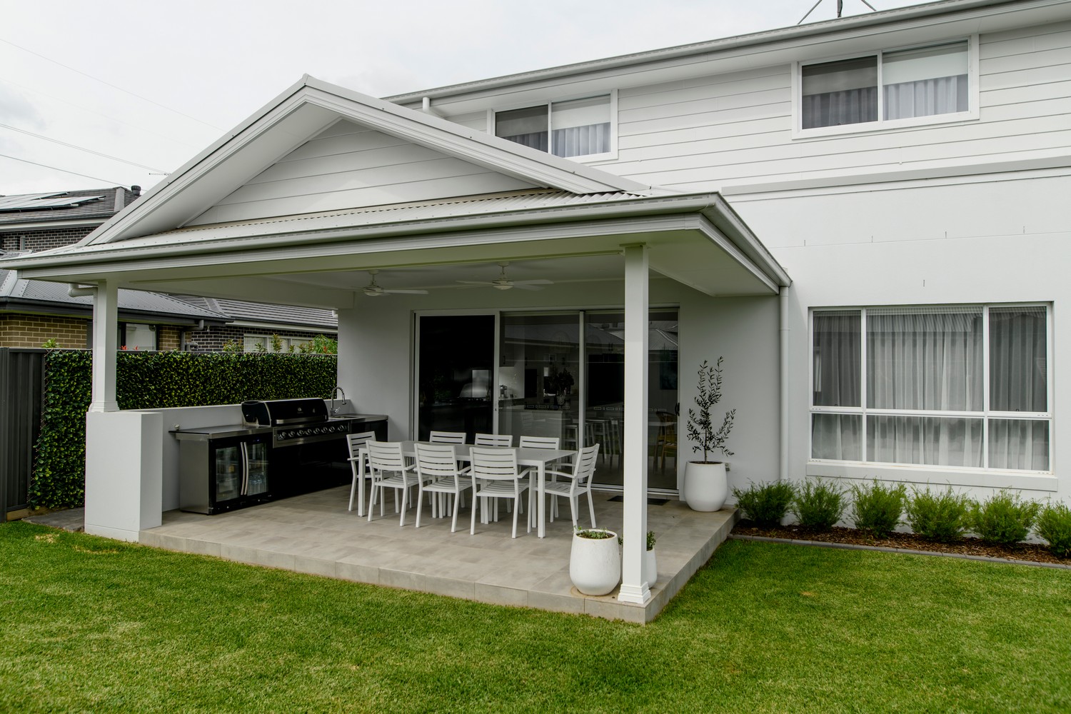 Modern outdoor patio with a concrete area, white dining set, barbecue, and greenery, featuring a sleek, minimalist house design.