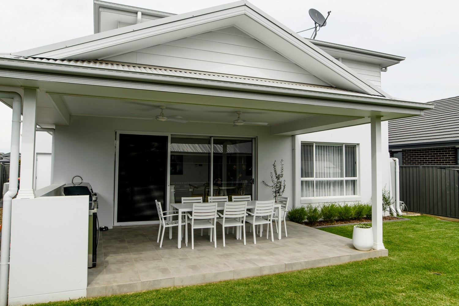 Contemporary outdoor dining area with a white table and chairs under a covered patio, surrounded by green lawn and modern home exterior.