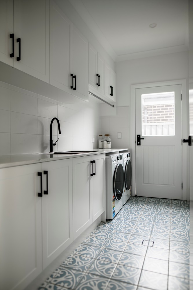 Bright laundry room featuring white cabinetry, a black tap, and dual washing machines, complemented by a decorative blue floor tile.