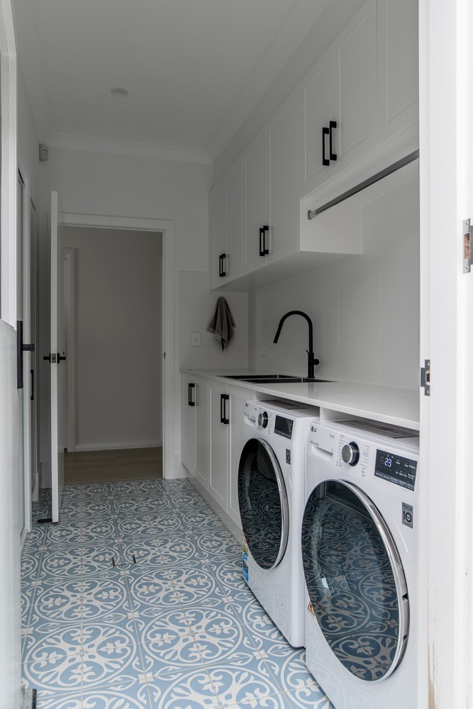 Bright laundry room featuring two white washing machines, sleek black fixtures, white cabinetry, and patterned blue floor tiles.