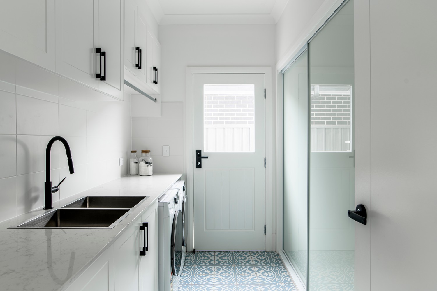 Bright laundry room featuring sleek white cabinetry, a modern sink, and patterned blue tiles, with a glass door allowing natural light.