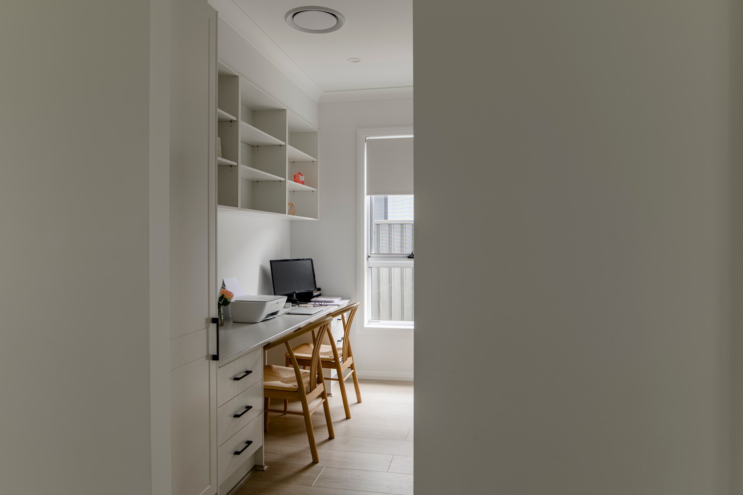 Bright home office with white walls and wooden flooring, featuring a desk with a computer and two chairs, shelves above for storage.