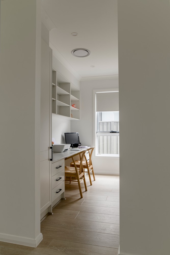 Bright home office with a white desk and wooden chair, featuring shelves above and natural light filtering through a window.