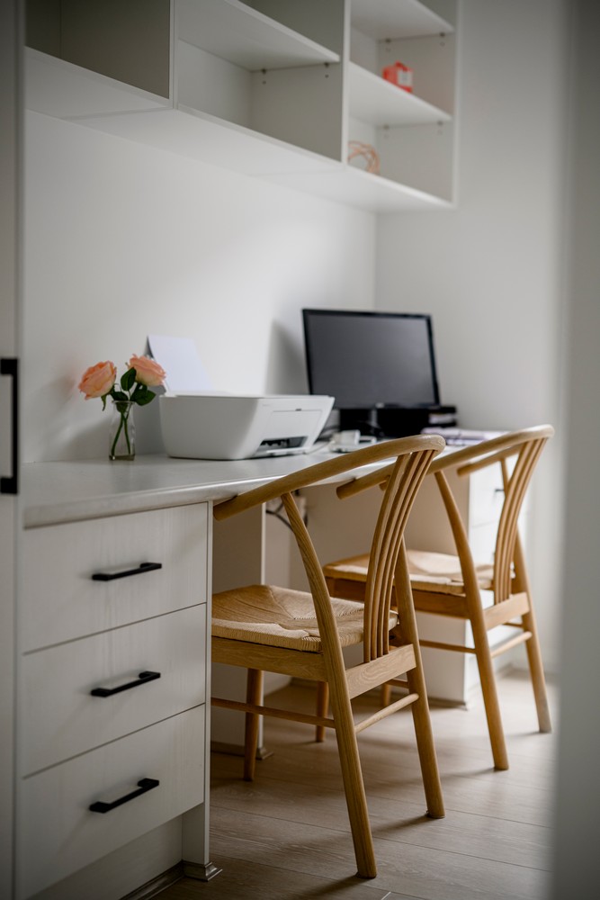 A modern home office with a light wooden desk, two rattan chairs, a computer, and a vase of pink roses against a neutral backdrop.