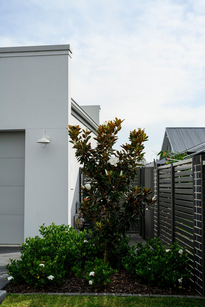 Bright exterior of a modern home featuring a magnolia tree, neat green shrubs, and a contemporary wooden fence, under a cloudy sky.