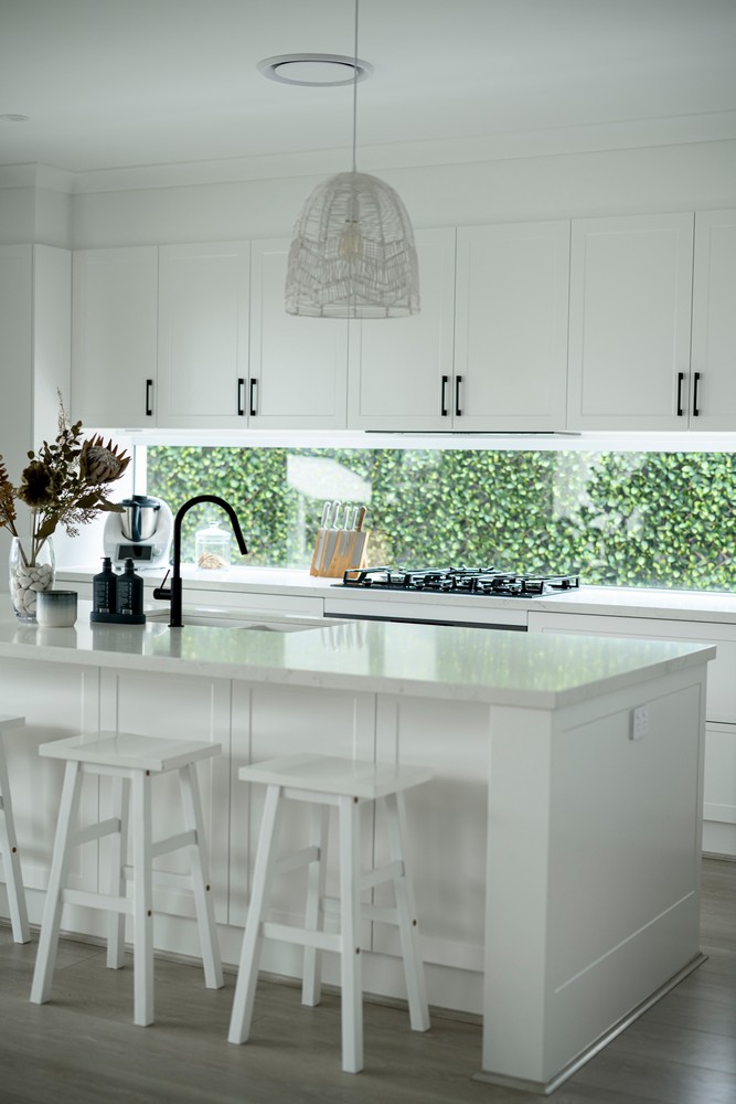 Bright, modern kitchen with a white island, bar stools, and a textured pendant light. Greenery visible through the large window.