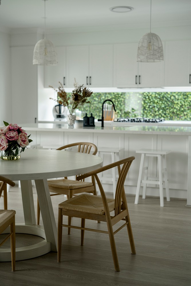 Bright, modern kitchen featuring a round wooden table with chairs, a floral centerpiece, and white cabinetry accented by green foliage outside.