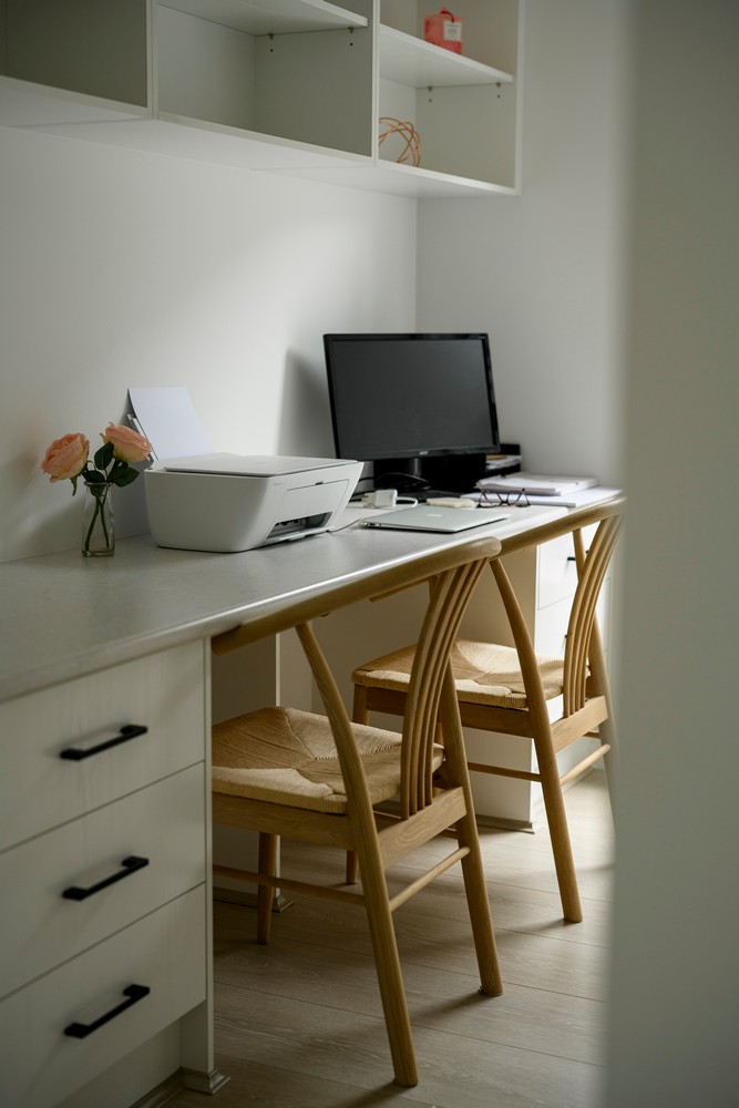 A minimalist home office featuring a light wooden desk with two chairs, a printer, and a computer monitor, complemented by a vase of roses.