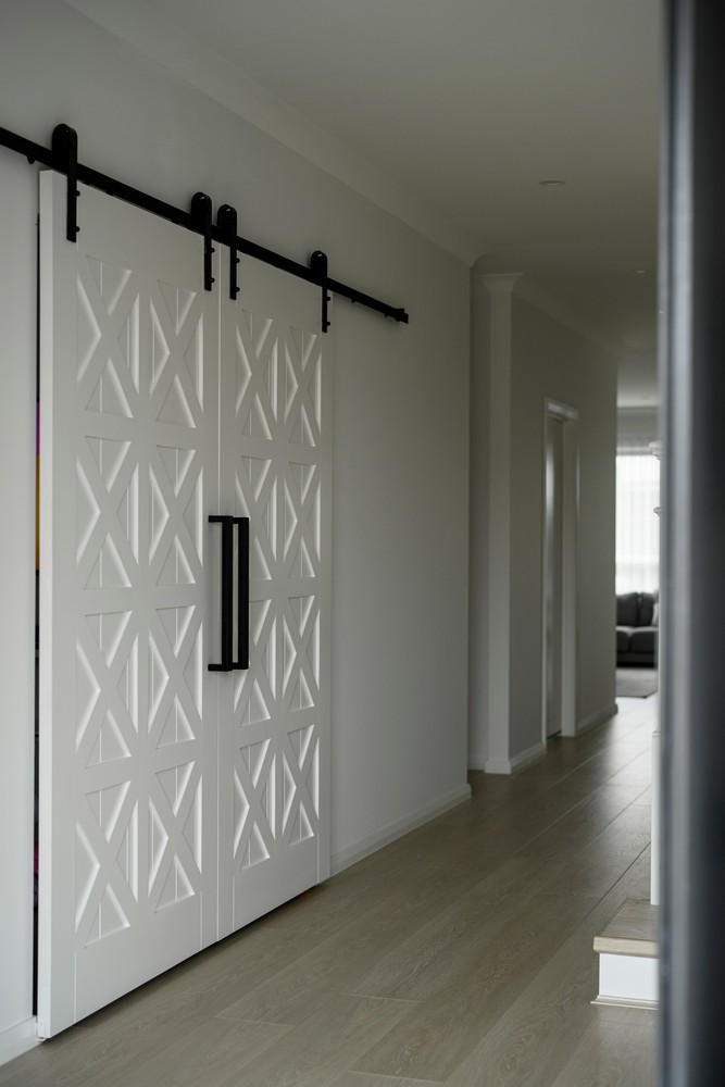 Stylish sliding white barn doors with an X-pattern design open to a modern hallway with light wooden flooring and grey walls.
