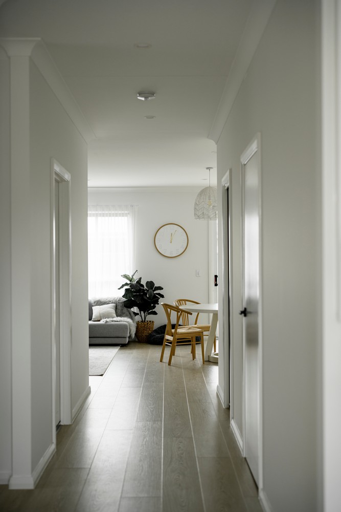 Bright hallway leading to a living area, featuring light wood flooring, green plants, a round wall clock, and a small dining set.