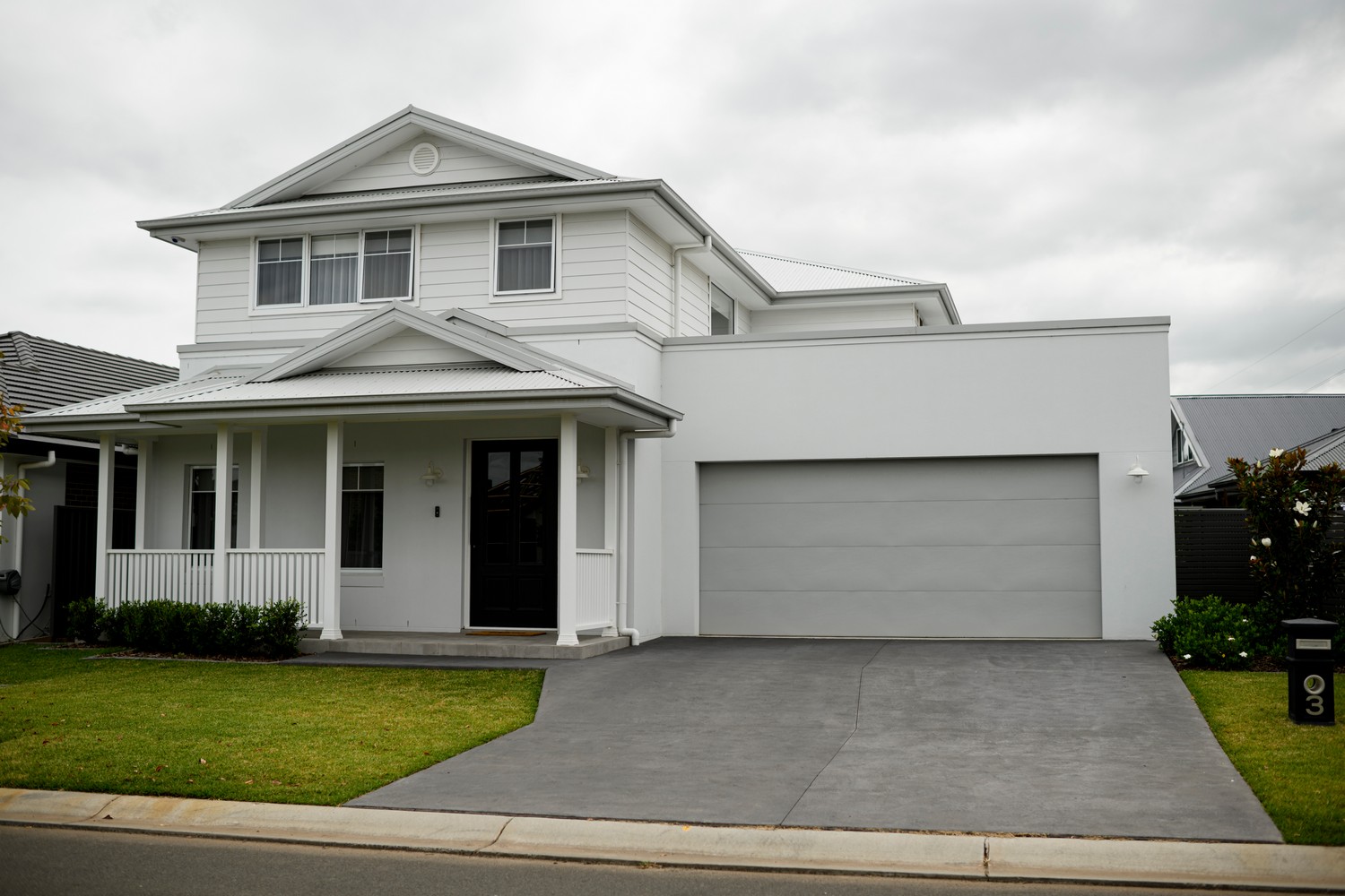 Modern two-storey house with a white facade, black front door, and grey garage. Well-maintained lawn and driveway. Overcast sky.