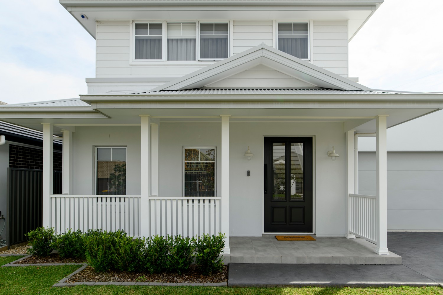 A modern two-storey home with white cladding, a dark front door, porch with railings, and neat greenery in the foreground.