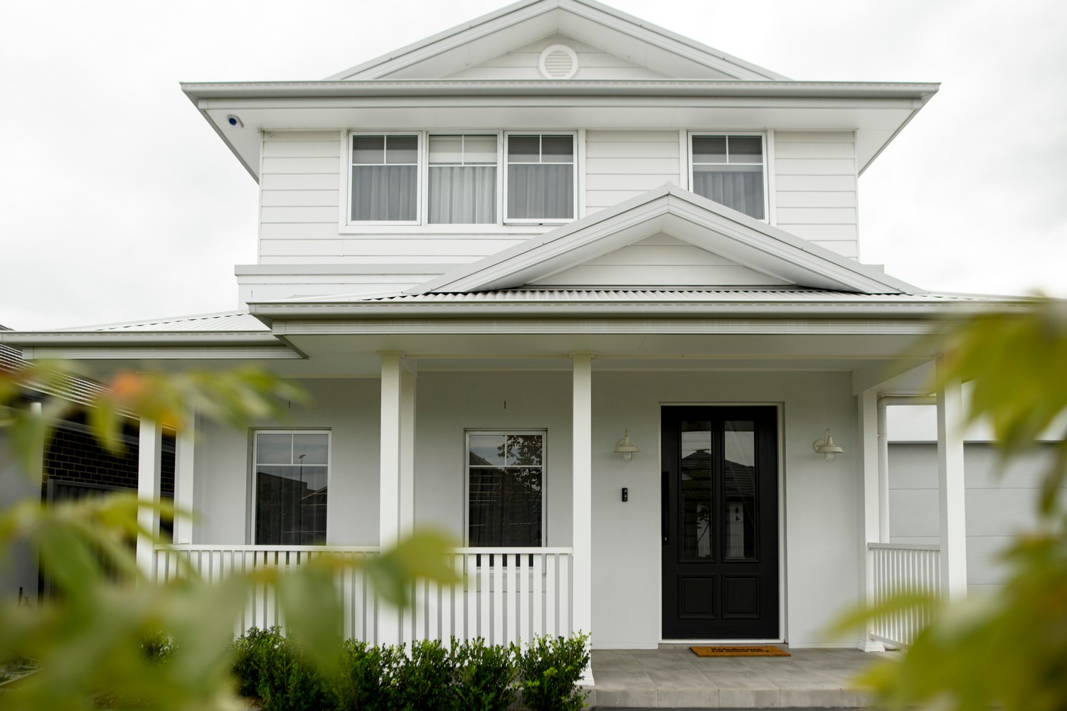 A modern two-storey white house with a black front door, large windows, and a welcoming porch, framed by green foliage.
