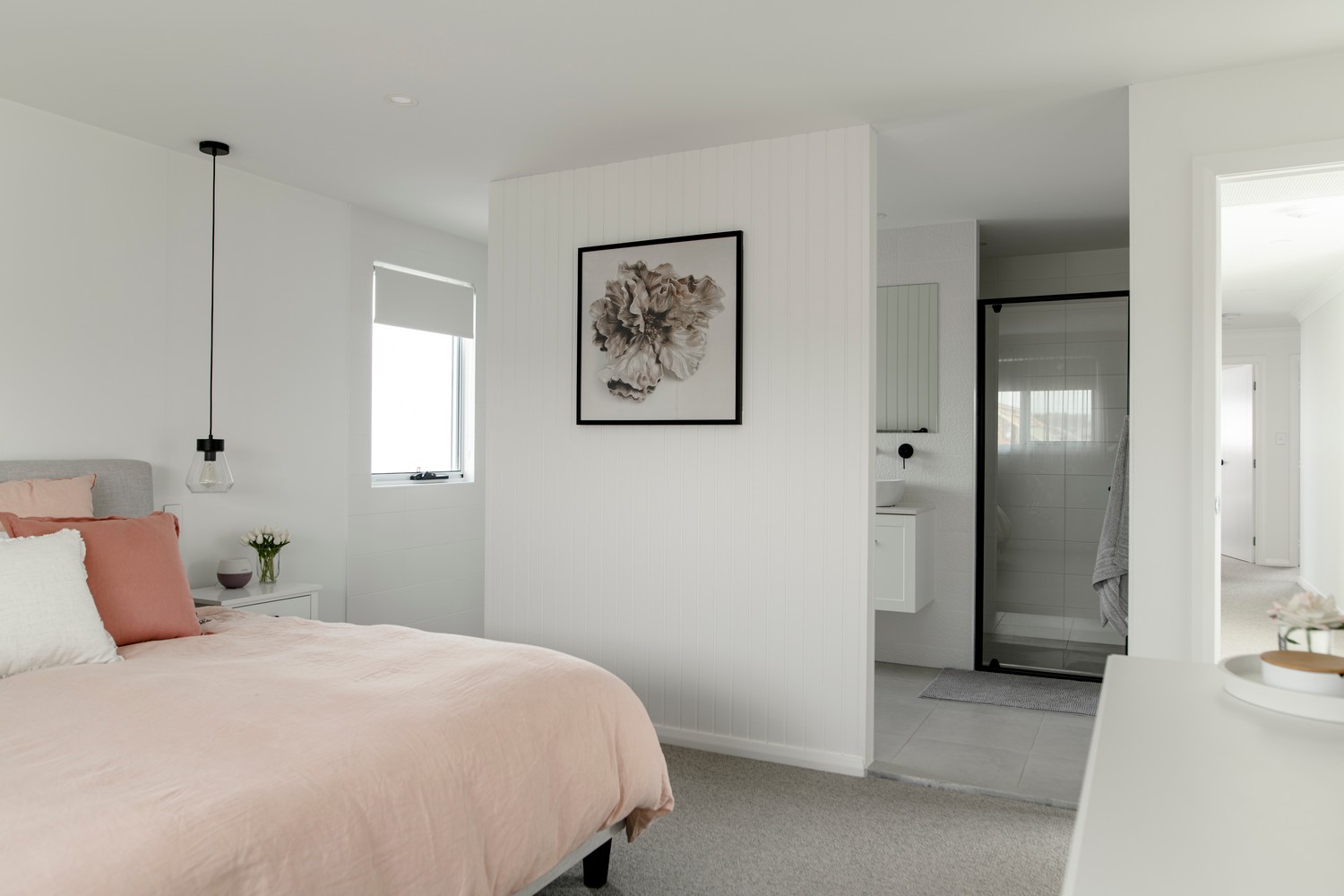 Cozy bedroom featuring a pink bedspread, a floral artwork, and an ensuite bathroom with a glass shower, set in soft neutral tones.