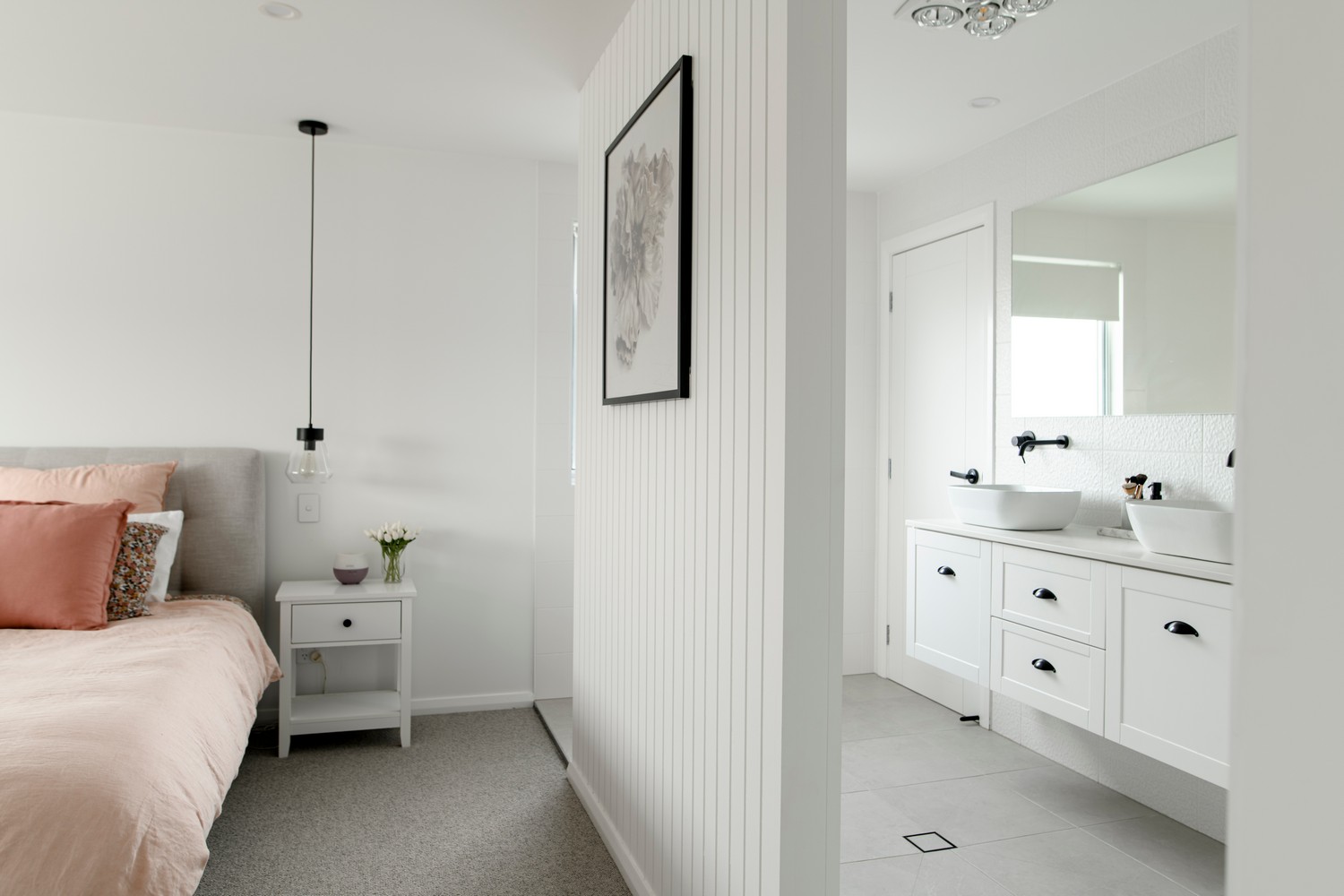 Elegant bedroom with a grey bed, soft pink bedding, and a nightstand. An adjoining bathroom features a sleek white vanity and modern decor.
