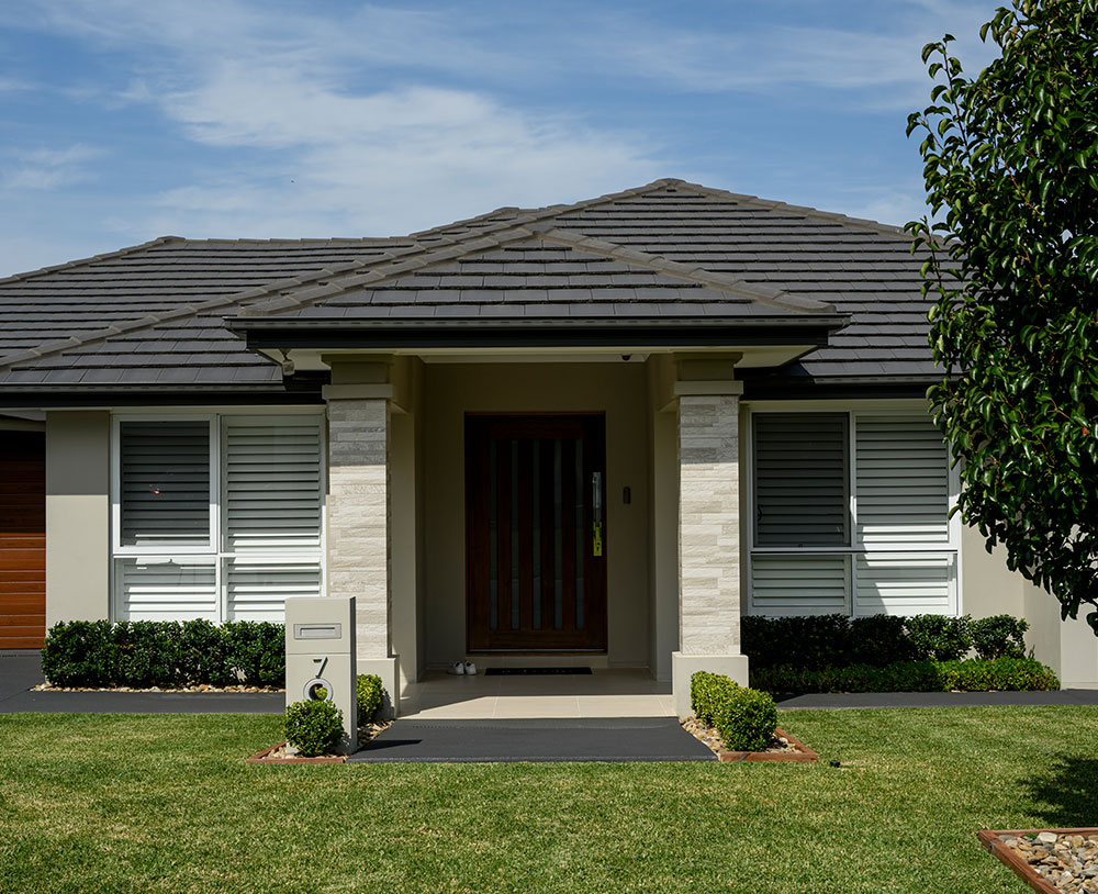 A modern home with a pitched roof, stone accents, and a front porch, surrounded by manicured lawns and hedges under a clear blue sky.