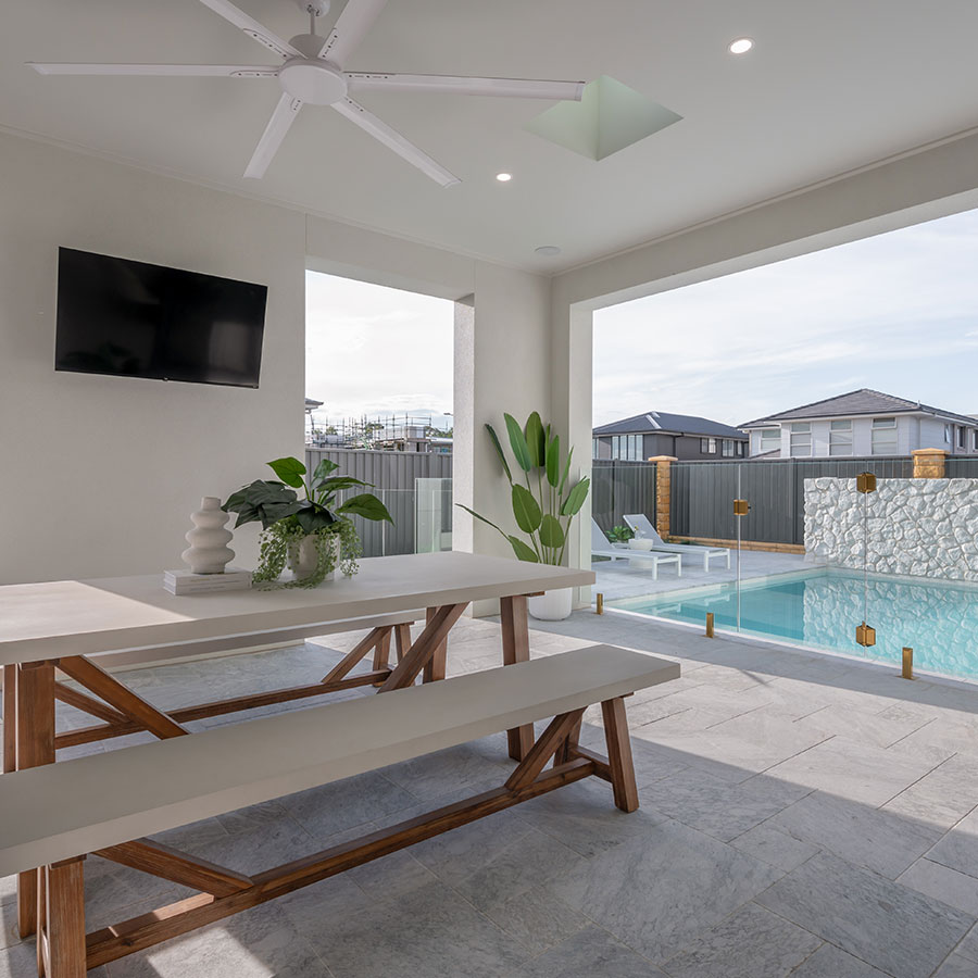 Bright outdoor space featuring a wooden dining table, planters, and a swimming pool, with modern architecture in the background.