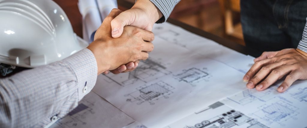 Two hands shake over architectural plans on a table, with a construction helmet nearby, symbolising collaboration in home design.